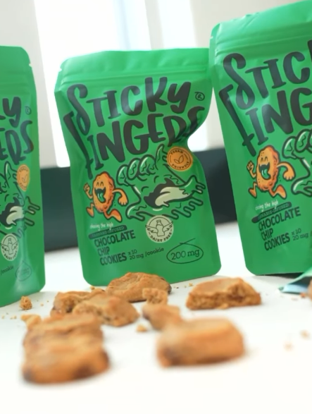 Three StickyFingers chocolate chip cookie packs with cookies scattered in front